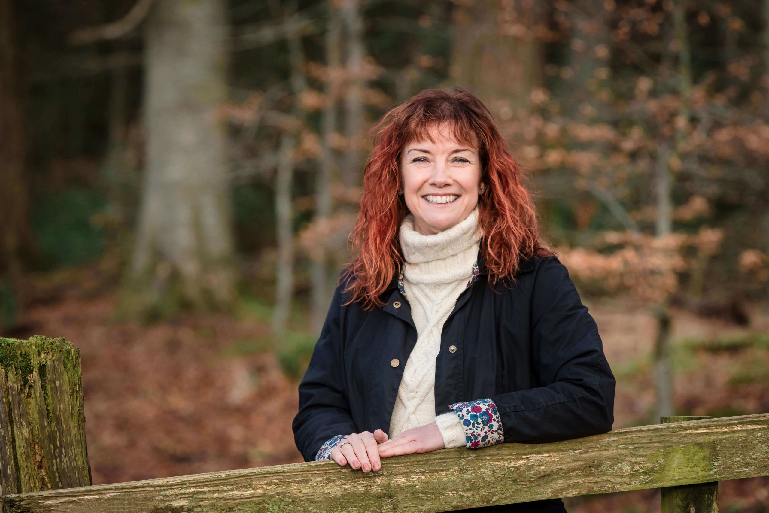 Woman in a navy jacket and cream jumper leaning on a wooden fence in autumn woodland